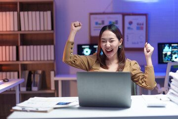 Asian female entrepreneur raising arms in excitement after achieving a business target using laptop computer