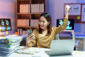 Asian female entrepreneur reading good news on mobile phone and raising arm in modern workplace