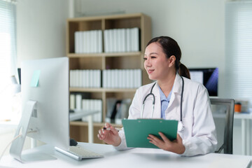 Asian female doctor holding a clipboard while working on a computer in a modern clinic, providing patient care and conducting online consultations