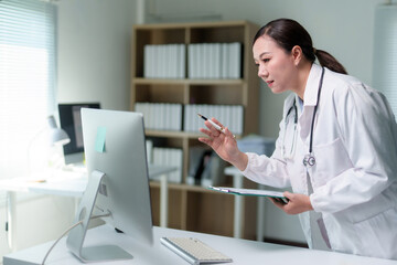 Asian female doctor wearing lab coat and stethoscope using desktop computer and holding clipboard, analyzing patient data in modern medical office