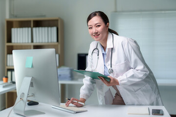 Asian female doctor wearing lab coat and stethoscope using computer and holding medical record in hospital office