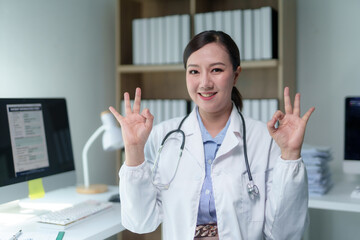 Asian female doctor wearing lab coat and stethoscope showing ok sign with both hands, standing in her office