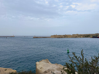 Coastal view of calm sea with rocky shoreline and distant harbor under a partly cloudy sky.