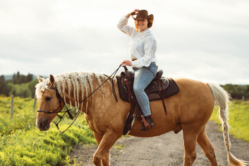 A woman, wearing a cowboy hat, is riding her horse down a dirt road
