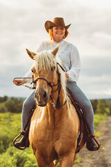 A woman, wearing a cowboy hat, is riding her horse down a dirt road