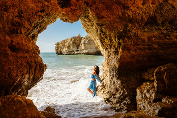Beautiful woman in blue dress posing below the arch in a stunning beach cave- Albufeira, Portugal