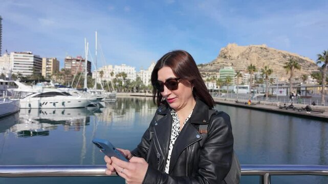 Young woman wearing sunglasses and a black leather jacket using smartphone in alicante port, with luxury yachts and mount benacantil in the background