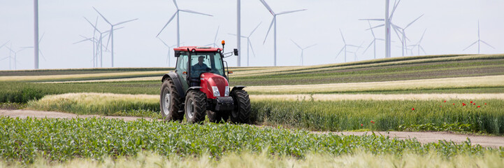 Obraz premium Scarlet tractor traverses verdant fields under towering windmills; celebrates Earth Day, sustainable farming, eco-friendly agriculture revolution