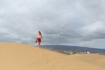 A woman in a white top and plaid skirt walks gracefully atop a sand dune, with distant mountains and buildings under cloudy skies.