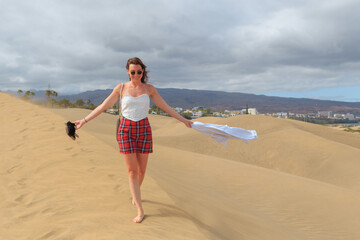 A woman strolls across golden sand dunes, wearing a light outfit and sunglasses, enjoying a breezy day against a mountainous backdrop.