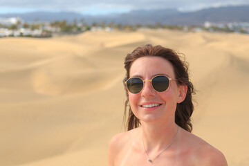 A woman in sunglasses smiles in a golden desert, enjoying the sun with scenic sand dunes behind her, capturing a joyful moment.