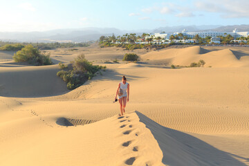 A traveler strides across golden sand dunes in Fuerteventura, enjoying the sunlight and serene landscape in the Canary Islands.