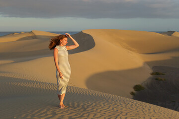 A woman strolls barefoot across golden sand dunes, enjoying the warm glow of the setting sun and gentle breeze in a tranquil desert landscape.