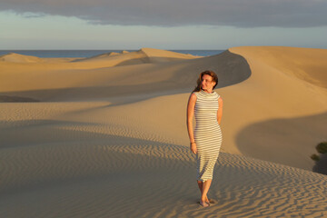 A woman strolls elegantly among golden sand dunes as the sun sets, casting long shadows across the landscape by the ocean.