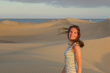 A young woman with flowing hair smiles while standing on soft sand dunes during sunset.