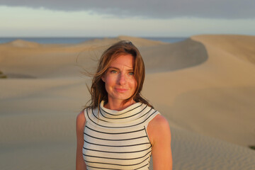 A woman with long hair poses in desert sand dunes as the sun sets, creating a tranquil and scenic atmosphere with a beach in the background.