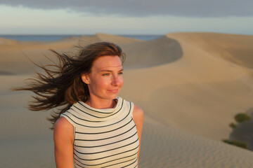A woman gazes thoughtfully over vast, sandy dunes as the sun begins to set, casting a warm glow over the landscape and enhancing her features.