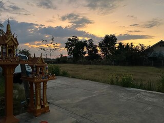 A serene image of a small Buddhist shrine nestled at the end of a quiet driveway in a village in...