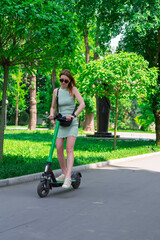 Modern urban mobility. A young stylish woman in a mint dress and sunglasses rides a green electric scooter in a city park on a summer day. Alternative transportation and modern technology