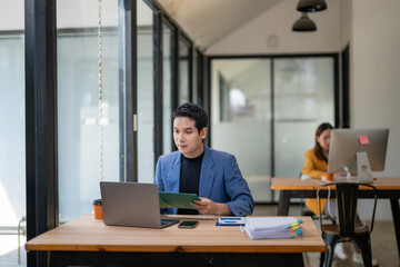 A man is sitting at a desk with a laptop and a stack of papers