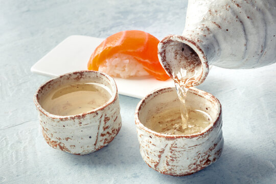 Sake poured into ochoko, traditional cup, with salmon nigiri in the background, traditional Japanese food and drink