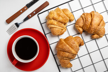 Knife and fork with freshly baked croissant on a cooling tray and a cup of coffee with a white background. Top view