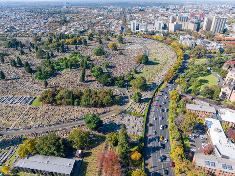 Fototapeta Aerial view of a busy city road winding around a cemetery towards high-rise buildings