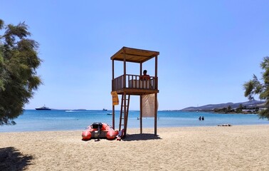 Photo of a peaceful summer view of Psaraliki beach in Antiparos island, Greece, with a lifeguard tower, clear turquoise waters, distant yachts, and swimmers enjoying the calm Aegean sea.