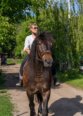 Rustic farm scene featuring a woman on a brown horse near a dirt path. Emphasizes tranquility,...