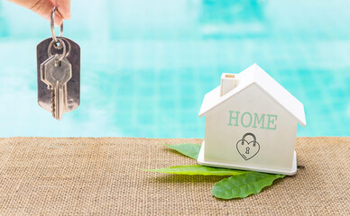 House key in girl hand with wooden house model on swimming pool edge, Green house, environmental concept, eco house