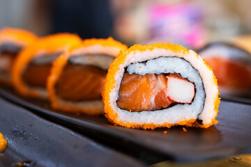 Sushi roll with salmon, crab stick, and rice, coated with orange tobiko, served on a black tray in a Japanese restaurant setting.