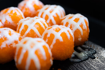 Sushi balls covered in bright orange tobiko and drizzled with white mayonnaise on a black tray, highlighting texture and vibrant color.