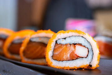Sushi roll with salmon, crab stick, and rice, coated with orange tobiko, served on a black tray in a Japanese restaurant setting.