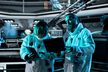 Teamworking astronauts doing brainstorming to confirm suit integrity using laptop. Orbital station...