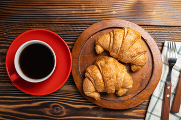 Fork and spoon on a napkin with a freshly baked croissant on a wooden tray with a cup of coffee on a wooden background. Top view