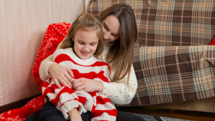 Happy family having fun at home for Christmas. Mother tickles her daughter sitting on the floor. Festive cozy interior