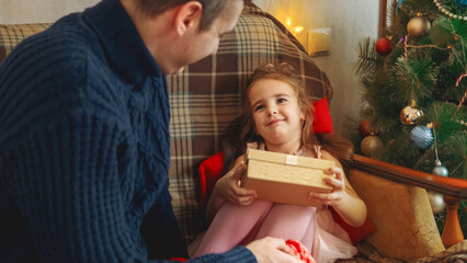 Dad is giving a gift box to his beautiful little daughter. Near the Christmas tree at home