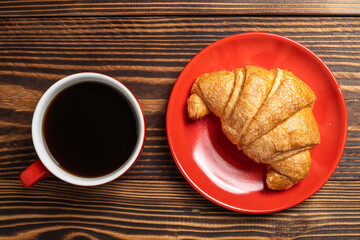 Freshly baked croissant on a red plate with a cup of coffee on a wooden background. Top view