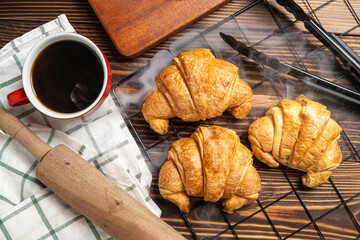 Rolling pin and tongs with freshly baked croissant on a cooling tray and a cup of coffee with a...