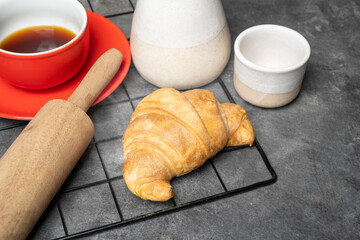 Freshly baked croissant and a cup of coffee with a rolling pin on a cooling tray on the table