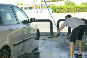 Rear view of adult man focused on car washing task using hose with water spray at urban car wash. Masculinity, care for possessions, focus on task, and modern man image in everyday context.