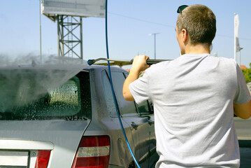 Man washing silver-gray passenger car with high-pressure hose at self-service station on summer day. Cleaning vehicle exterior, car maintenance routine, DIY car care and everyday cleanliness