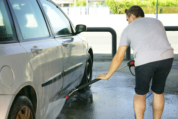 A young man bends over and washes the underside of a silver-gray passenger car with a high-pressure water gun at a self-service car wash in an open-type bay. Self-cleaning of personal transport