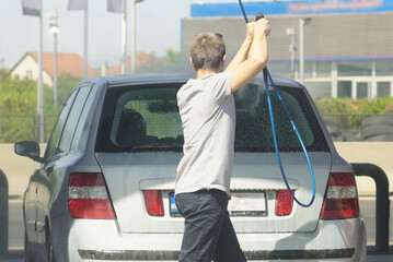 Young man washing rear of a passenger car with high-pressure water hose. Masculine responsibility, vehicle upkeep, urban environment, person in casual moment and self-service lifestyle snapshot.