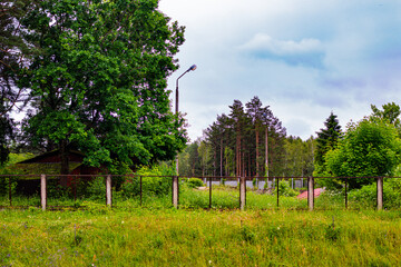 Old fenced compound with overgrown grass, trees, and an abandoned building, surrounded by dense...