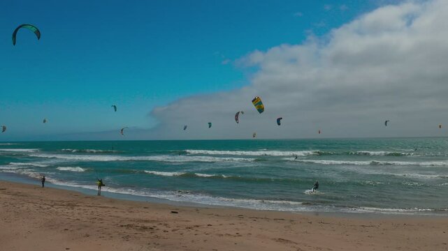 Kitesurfers ride ocean waves near Pacifica, California, along Highway 1. Colorful kites fill the cloudy sky over the turquoise sea in this action-packed coastal scene