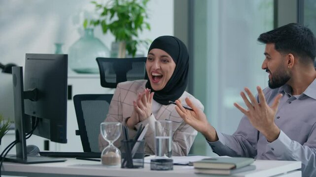 Two Islamic Arabian Muslim business team colleagues celebrate online pc success at office table businesswoman in hijab with businessman happy amazed computer work achievement victory high five gesture
