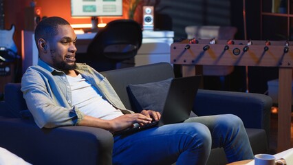 Adult guy typing on laptop laying on couch in stylish apartment, engaging in freelancing and online...