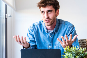 Confident young man sitting at desk with laptop, smiling and speaking directly to the camera....