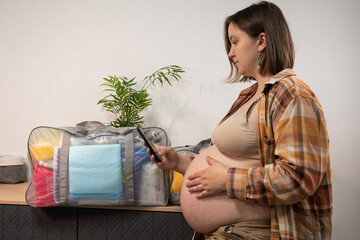 Pregnant woman at 9 months of pregnancy collects a bag in the hospital. Future mom preparing and planning clothes to the newborn baby. Checking the list using the phone